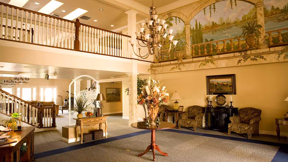 Spacious elegant lobby with a central table holding a floral arrangement, chandelier, seating area, and a painted wall mural.