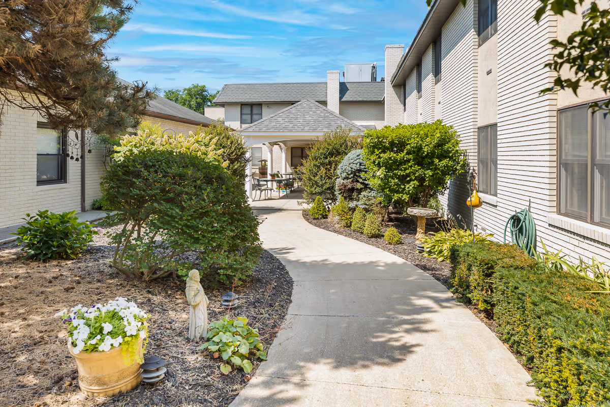 Curved concrete walkway through a landscaped courtyard between light brick buildings with shrubs, potted flowers and a small statue.