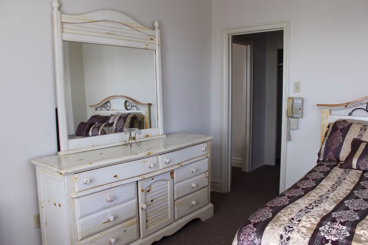A bedroom featuring a white distressed wooden dresser with a large mirror on top. The bed has a headboard with a metal design and is covered with a striped purple, beige, and brown patterned bedspread. There is a doorway leading to another room, and a wall-mounted telephone is visible next to the door.