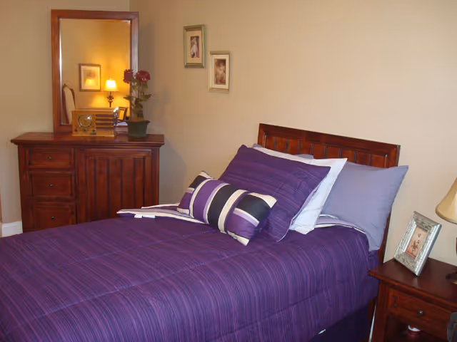 A bedroom with a wooden bed frame and a purple bedspread with matching pillows. There is a wooden dresser with a mirror and a vintage radio on top, along with a potted plant. Two framed pictures hang on the wall above the bed. A wooden nightstand with a lamp and a framed photo is next to the bed.