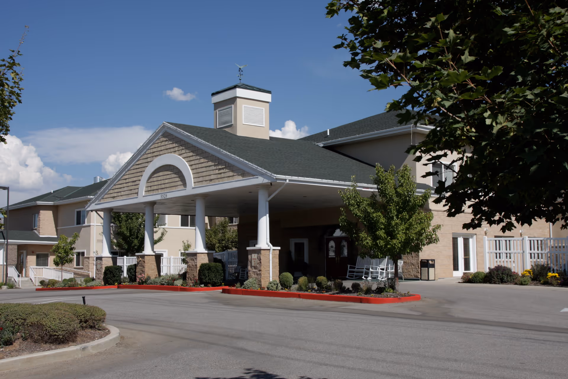 Exterior view of Legacy House Assisted Living of Ogden showing the main entrance with a covered driveway supported by white columns. The building has beige walls, a green roof, and landscaping with trees and shrubs under a clear blue sky.
