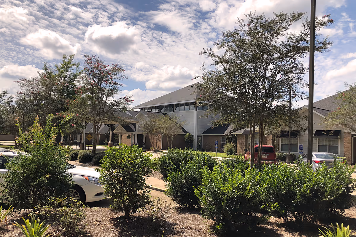 Exterior view of Oak Park Village at Slidell senior living facility showing a parking area with several cars, landscaped bushes and trees, and a partly cloudy sky.