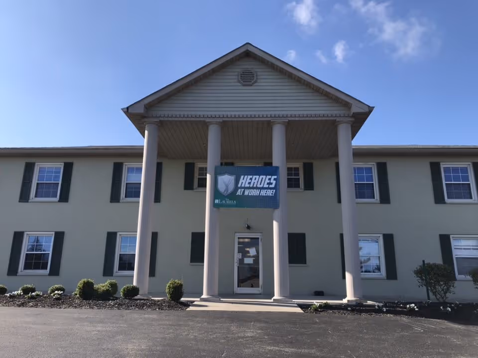 Front exterior of a two-story pale-green building with four columns and a "HEROES AT WORK HERE!" banner above the entrance.