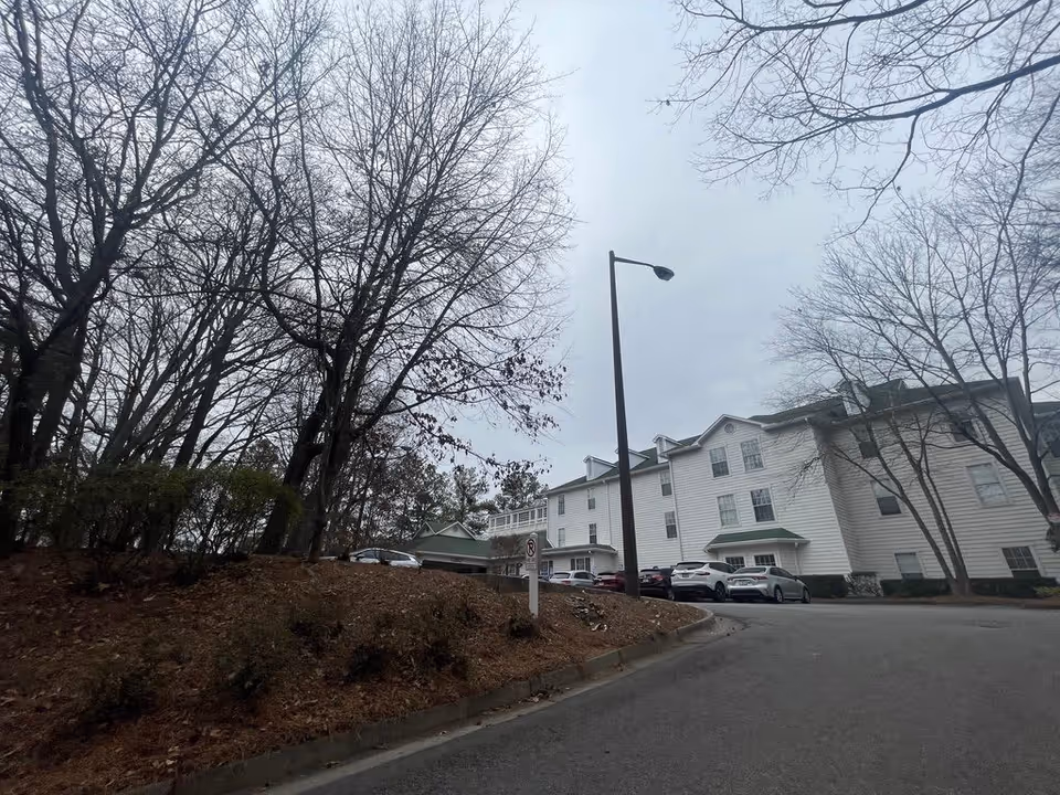 Exterior view of a three-story white senior living building with parked cars, leafless trees, and a lamppost on an overcast day.