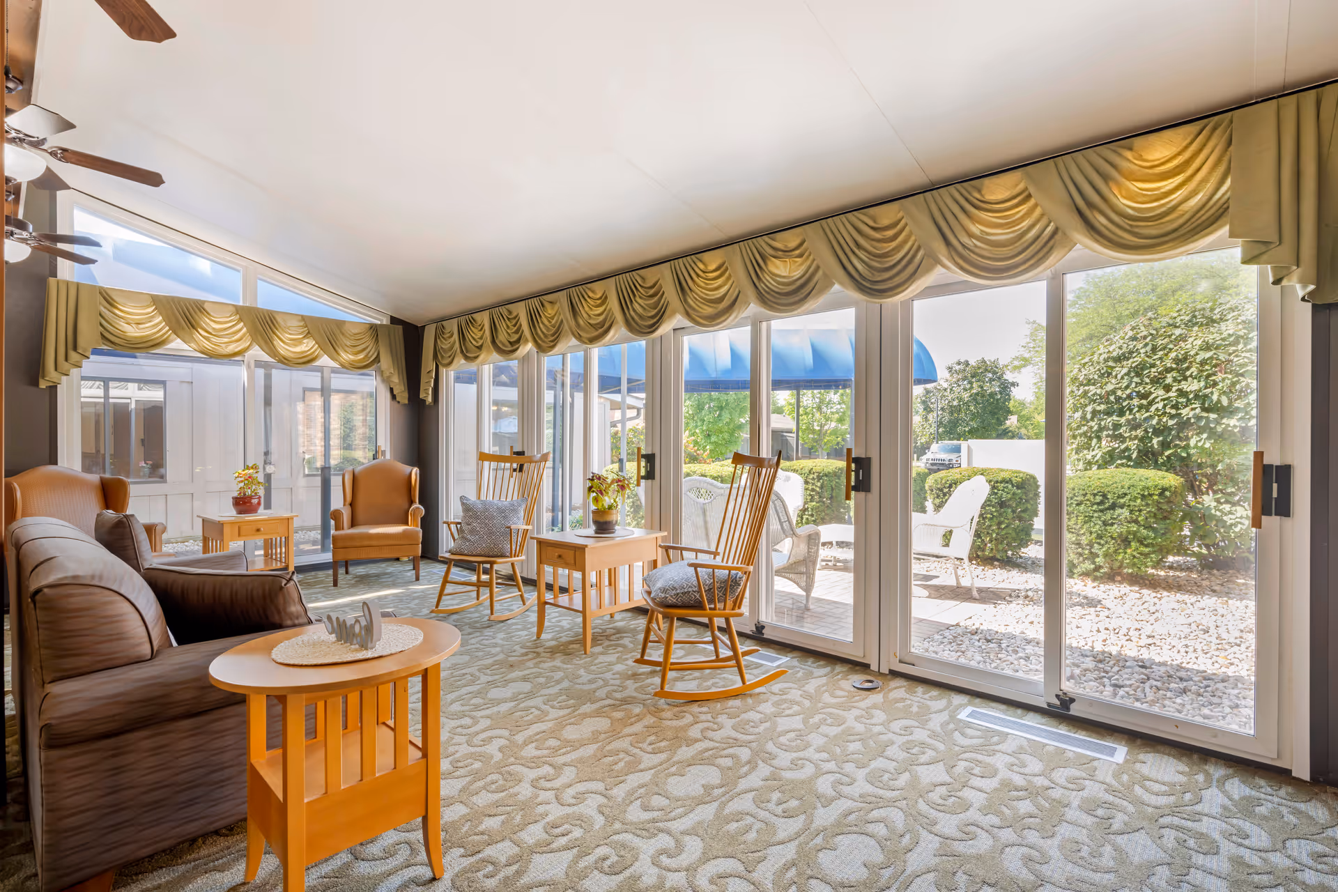 A bright and cozy living room area with large floor-to-ceiling windows and sliding glass doors that open to an outdoor patio. The room features a brown leather sofa, two wooden rocking chairs with cushions, two armchairs, and wooden side tables with potted plants. The windows are adorned with gold-colored valances, and the carpet has a light patterned design. Ceiling fans with lights are visible on the ceiling.