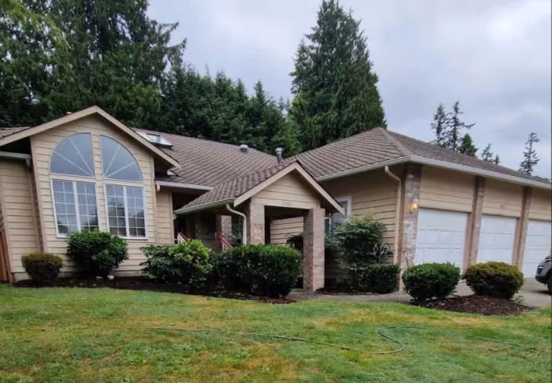 Exterior view of a single-story residential building with beige siding, a large arched window, a covered entrance, and a three-car garage. The building is surrounded by green bushes and a grassy lawn with tall trees in the background under a cloudy sky.