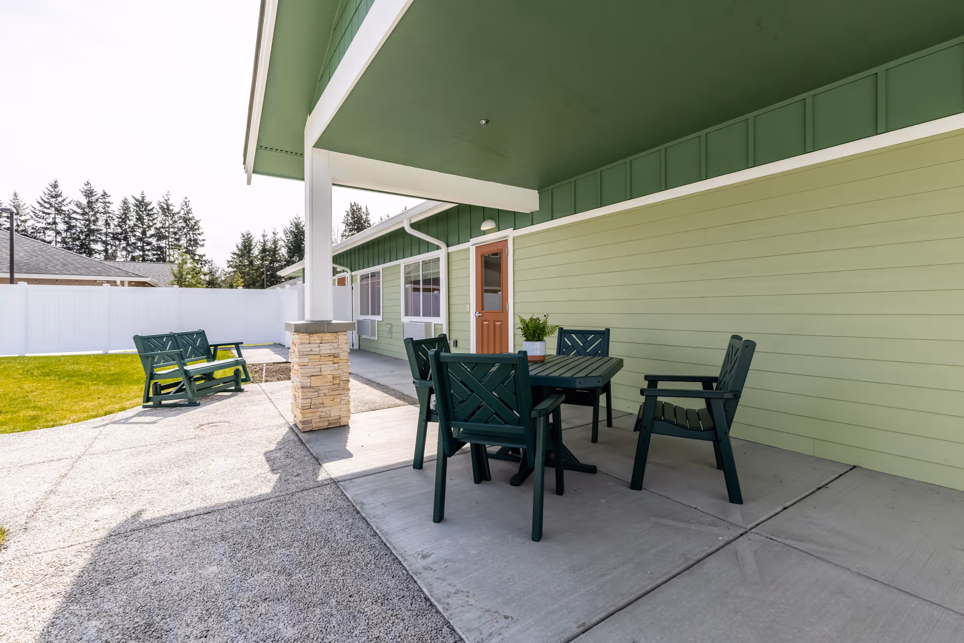 Outdoor patio area at Memory Haven with green plastic chairs and a table under a covered section attached to a light green building. There is a small potted plant on the table, a stone pillar supporting the roof, a white fence enclosing the grassy yard, and trees in the background.