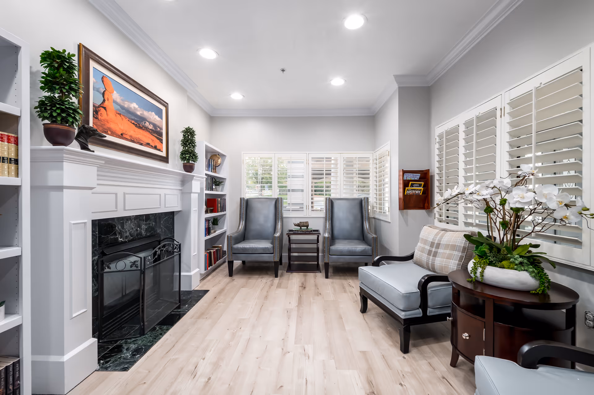 A bright and cozy living room area with light wood flooring, two gray armchairs with a small table between them, a light blue cushioned bench with a plaid pillow, and a round wooden side table holding a white orchid plant. The room features a white fireplace with a black marble hearth, built-in bookshelves filled with books and decorative items, and large windows with white plantation shutters. A framed landscape photograph hangs above the fireplace.