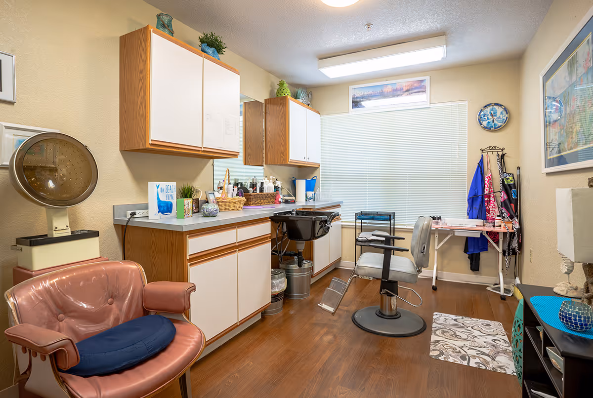 Interior of a salon room with a pink salon chair, a gray salon chair in front of a hair washing sink, wooden cabinets with white doors, a window with blinds, a small table with hairdressing tools, and various decorative items on the walls and shelves.
