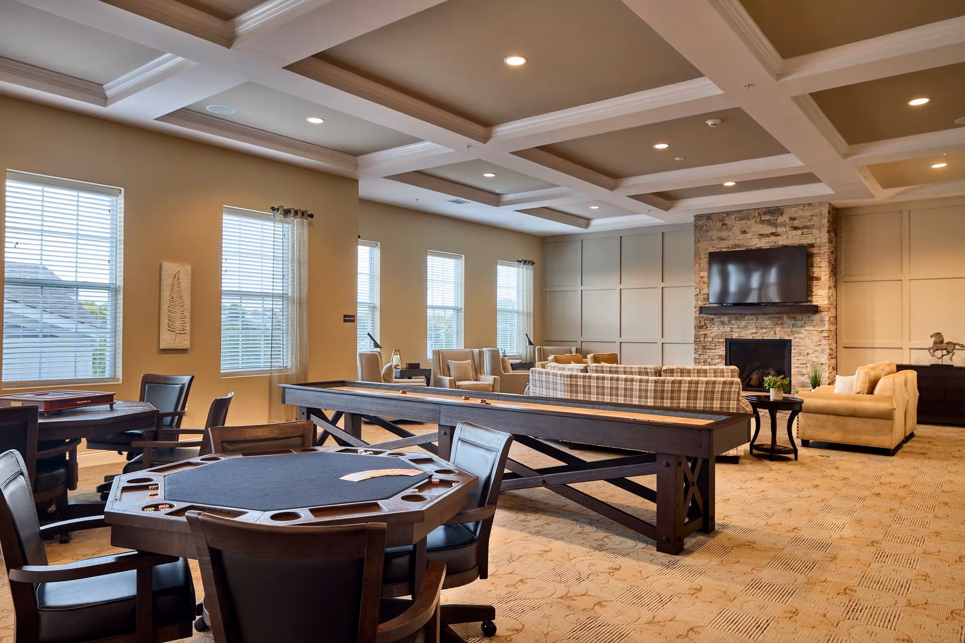 A spacious and well-lit common area in a senior living facility featuring a card table with chairs, a shuffleboard table, and a cozy seating area with sofas and armchairs arranged around a stone fireplace with a mounted flat-screen TV above it. Large windows with blinds allow natural light to fill the room, and the ceiling has decorative beams with recessed lighting.