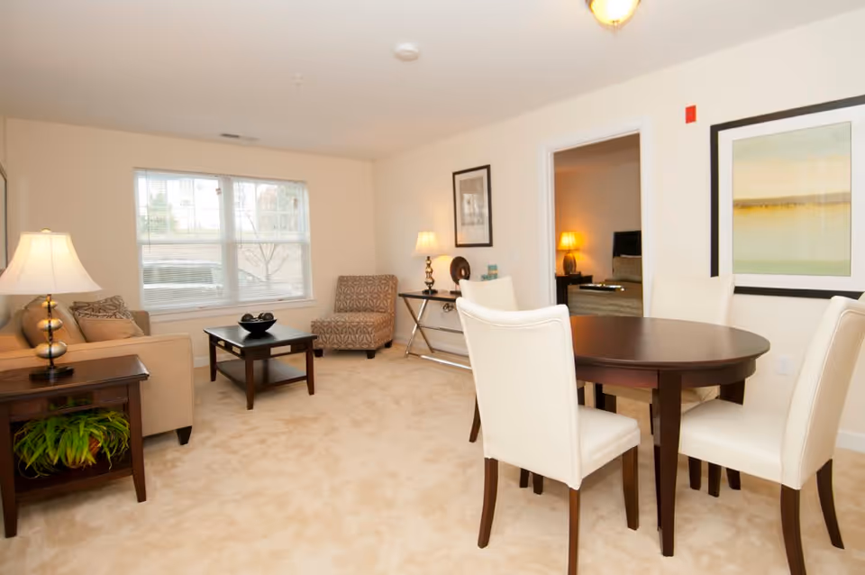 Bright furnished living and dining area with a round dark wood table and white chairs, sofa, armchair, coffee table and lamps.