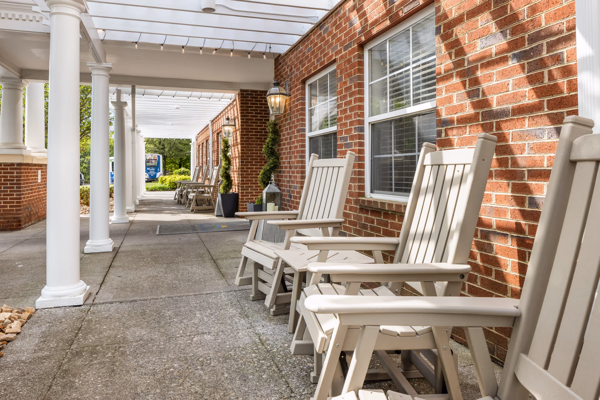 Outdoor covered walkway with white columns and beige rocking chairs lined up against a red brick wall with windows. There are decorative plants and lanterns along the wall, and string lights hanging from the white pergola overhead.