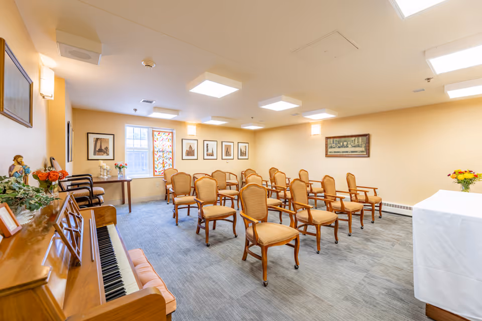 A small chapel or meeting room with rows of wooden chairs with cushioned seats arranged facing a white altar covered with a cloth. The room has beige walls, framed pictures on the walls, a piano on the left side with flowers and decorations on top, and a window with stained glass. The floor is carpeted in gray tones and the ceiling has several square light fixtures.