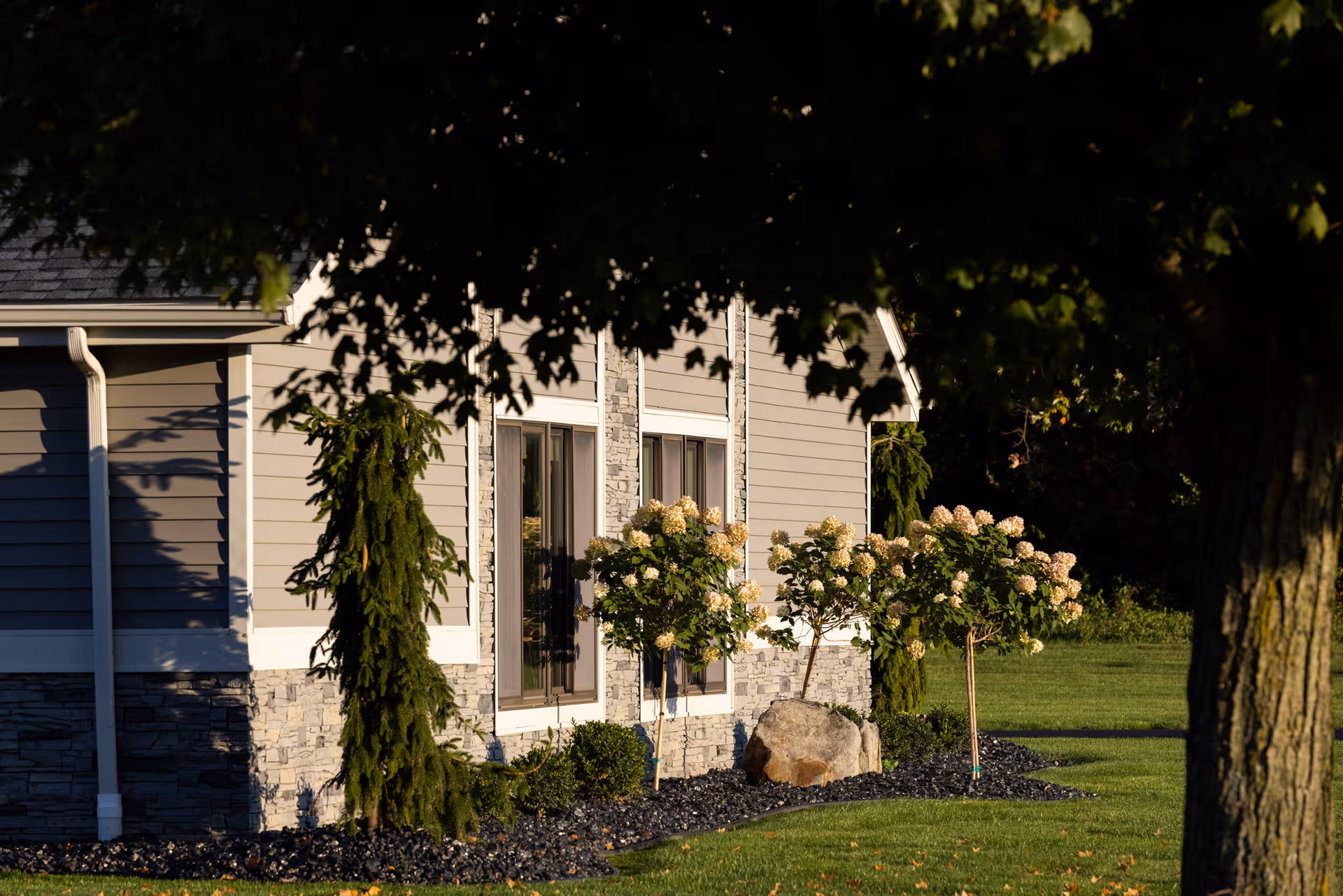 Exterior side view of a senior care facility building with beige siding and stone accents, surrounded by a landscaped garden with small trees, flowering shrubs, and a large rock, partially shaded by a large tree in the foreground.