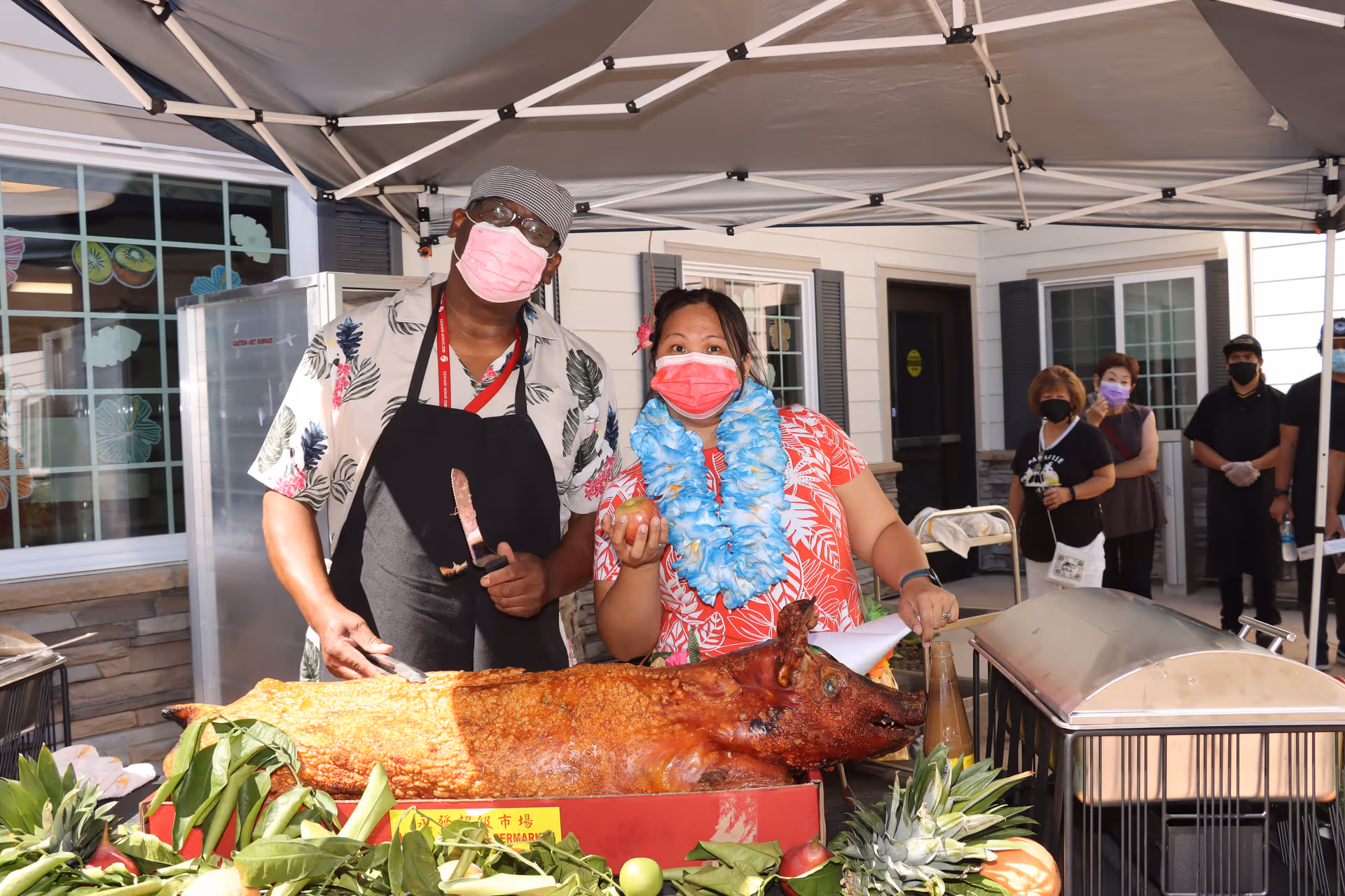 Two masked servers stand behind a table with a roasted whole pig and garnishes under a canopy outside a senior living building.