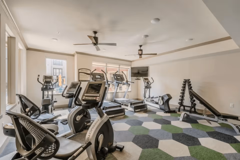 A fitness room with exercise equipment including stationary bikes, treadmills, an elliptical machine, a weight bench, and a rack of dumbbells. The room has large windows, ceiling fans, and a TV mounted on the wall. The floor is covered with a patterned carpet in shades of green, gray, and white.