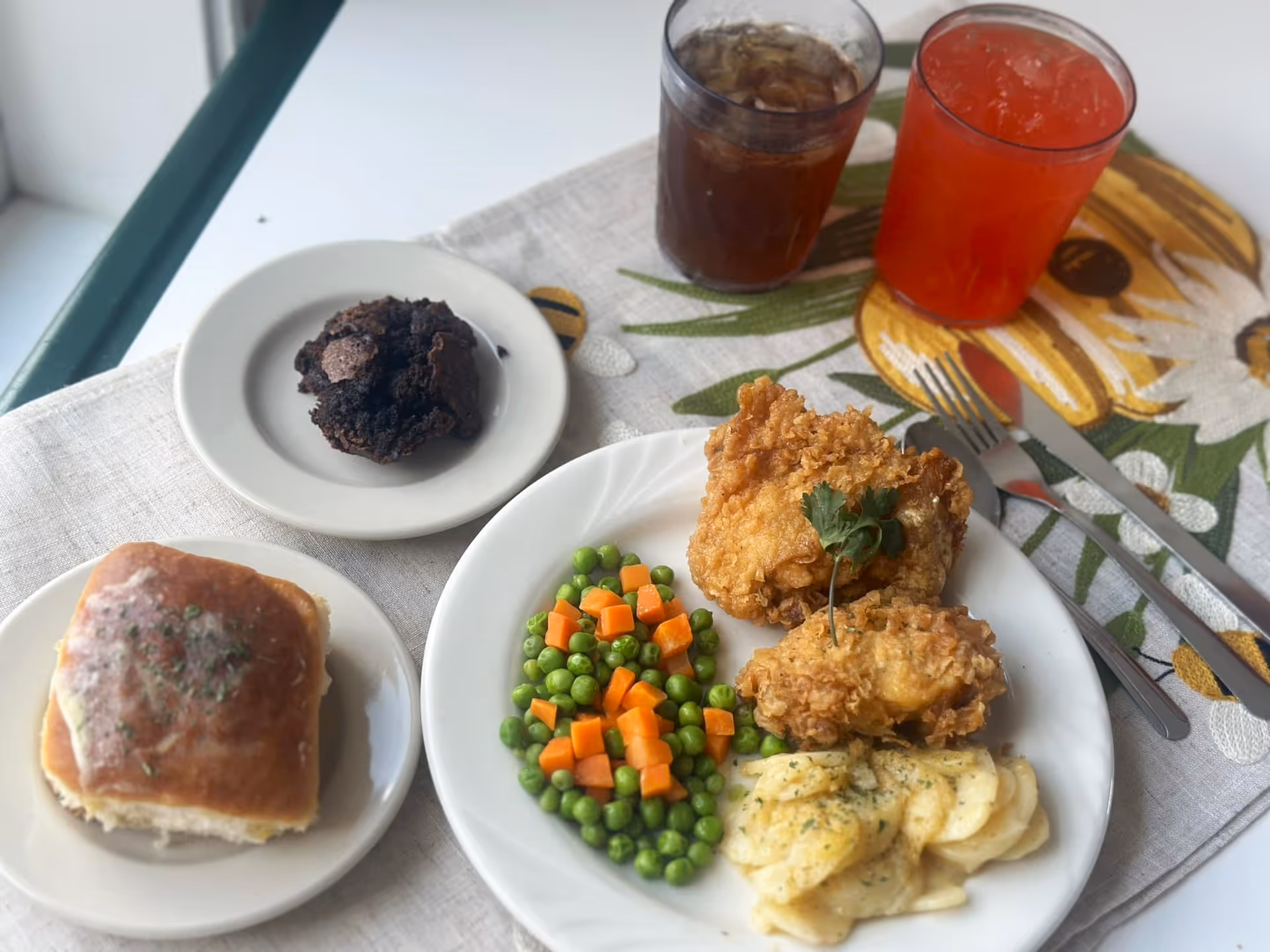 A meal served on a table with a plate containing fried chicken, peas and carrots, and scalloped potatoes. There is a dinner roll on a small plate, a chocolate muffin on another small plate, and two glasses of iced beverages, one dark and one red. The table is covered with a light-colored cloth and a floral placemat.