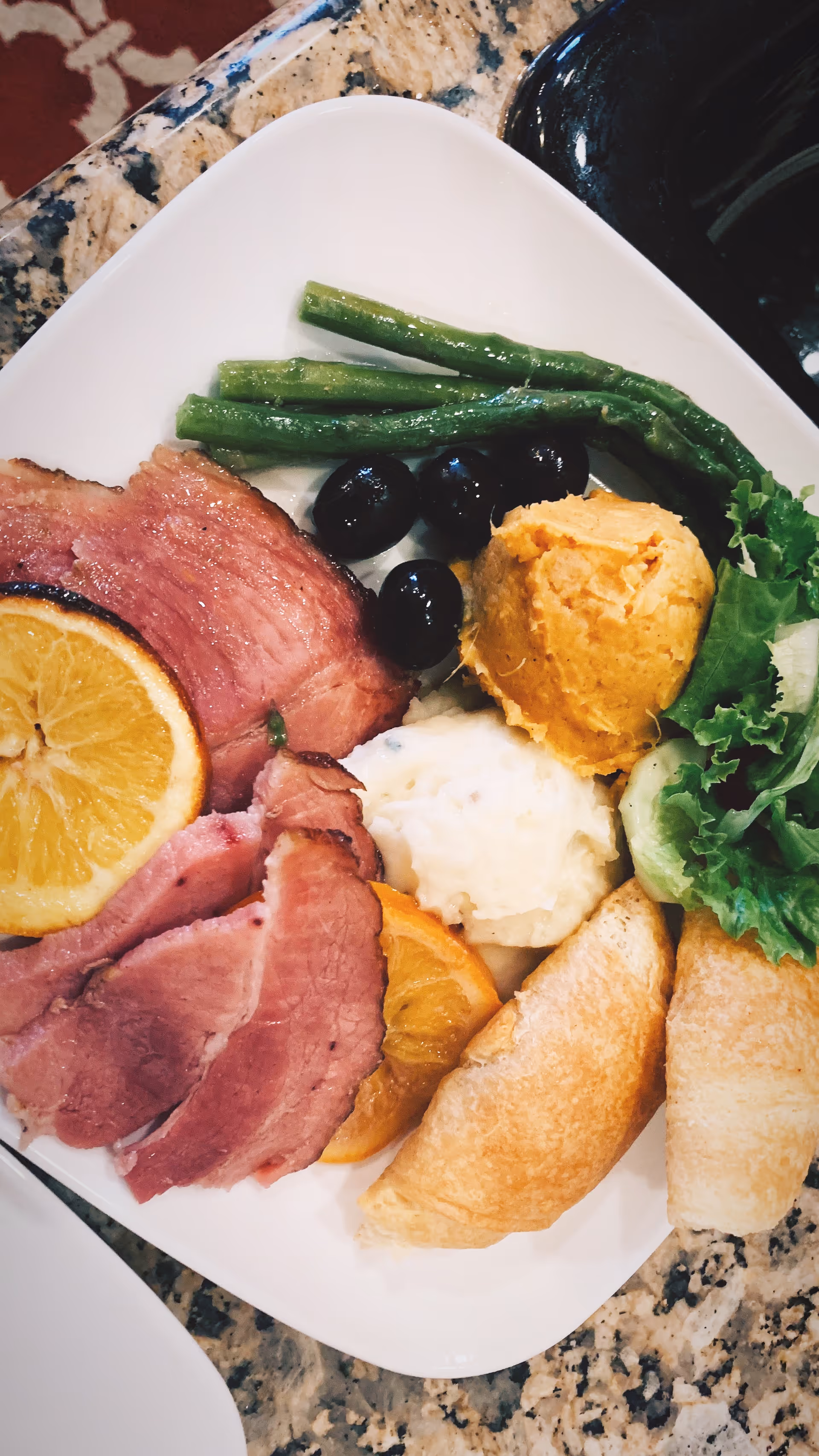 A plate of ham with orange slices, green beans, black olives, mashed potatoes, a scoop of sweet potato, salad greens and a croissant.