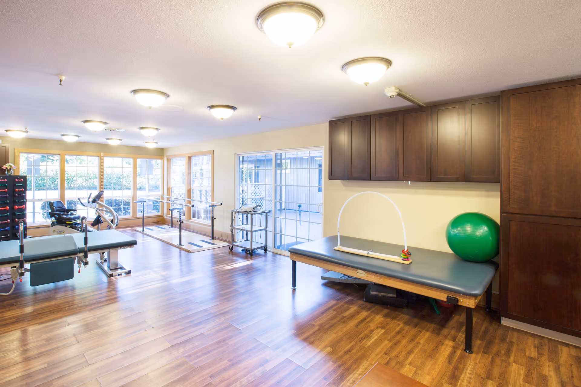 A bright rehabilitation room with wooden flooring, exercise equipment including parallel bars, a recumbent bike, therapy tables, a green exercise ball, and cabinets mounted on the wall. Large windows and a sliding glass door allow natural light to fill the space.