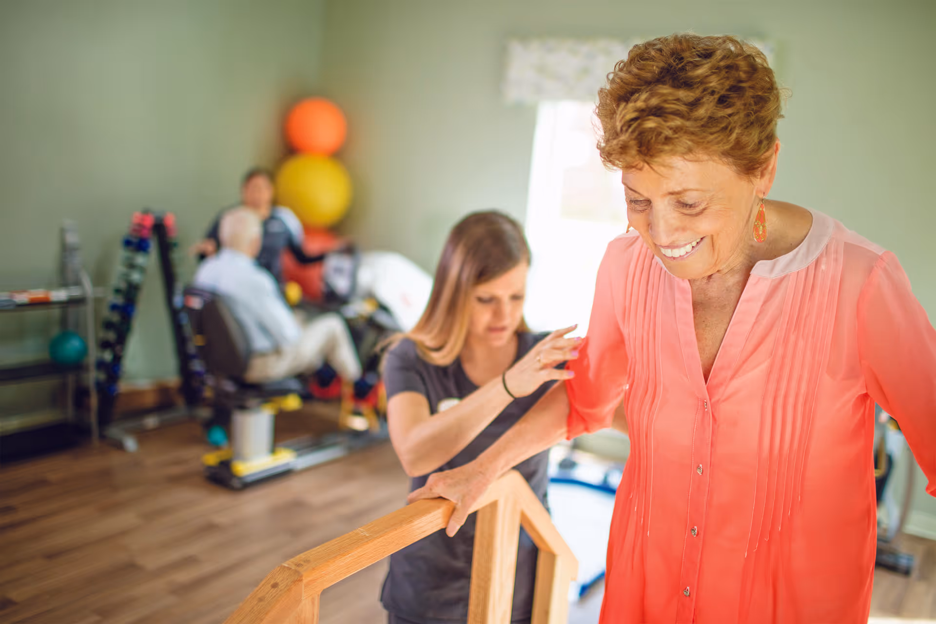 An elderly woman in a coral blouse is smiling while holding onto a wooden handrail as a younger woman assists her with physical therapy exercises in a room with exercise equipment and colorful therapy balls in the background.