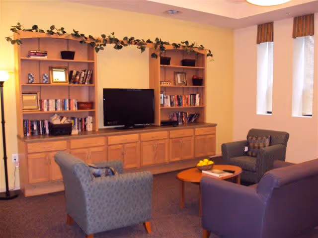A cozy living room area with three upholstered armchairs arranged around a wooden coffee table with a bowl of lemons and a book. Behind the chairs is a wooden entertainment center with shelves holding books, picture frames, and decorative items, and a flat-screen TV in the center. The walls are painted light yellow and there are three narrow windows with striped valances letting in natural light.