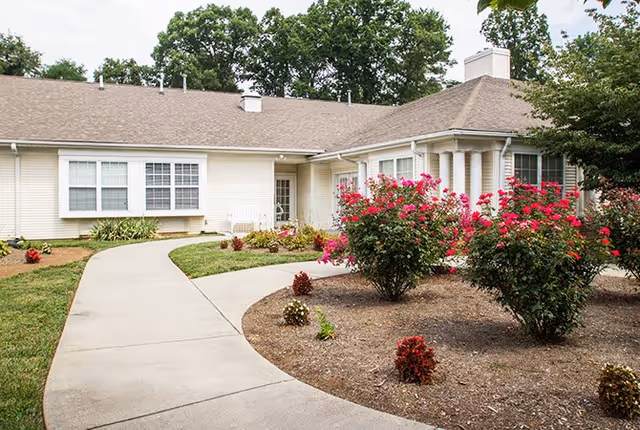 Exterior view of a single-story senior living facility building with beige siding and a brown shingled roof. A curved concrete walkway leads to the entrance, surrounded by landscaped flower beds with blooming pink and red flowers and small shrubs. Trees are visible in the background.
