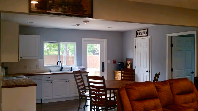 Interior view of a senior living facility kitchen and dining area with white cabinets, a wooden dining table with chairs, a brown sofa in the foreground, a window and a glass door leading outside, and a pantry door with a sign above it.