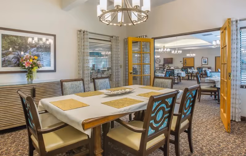 A dining room with a rectangular table covered with a white tablecloth and six chairs with patterned backs. The room has carpeted floors, a sideboard with a vase of flowers, and a framed picture on the wall. French doors open to a larger dining area with multiple tables and chairs, chandeliers, and windows with blinds and curtains.