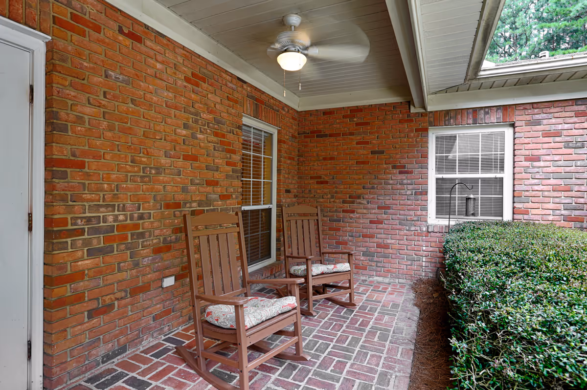 Covered brick porch with two wooden rocking chairs, a ceiling fan, windows, and hedges.