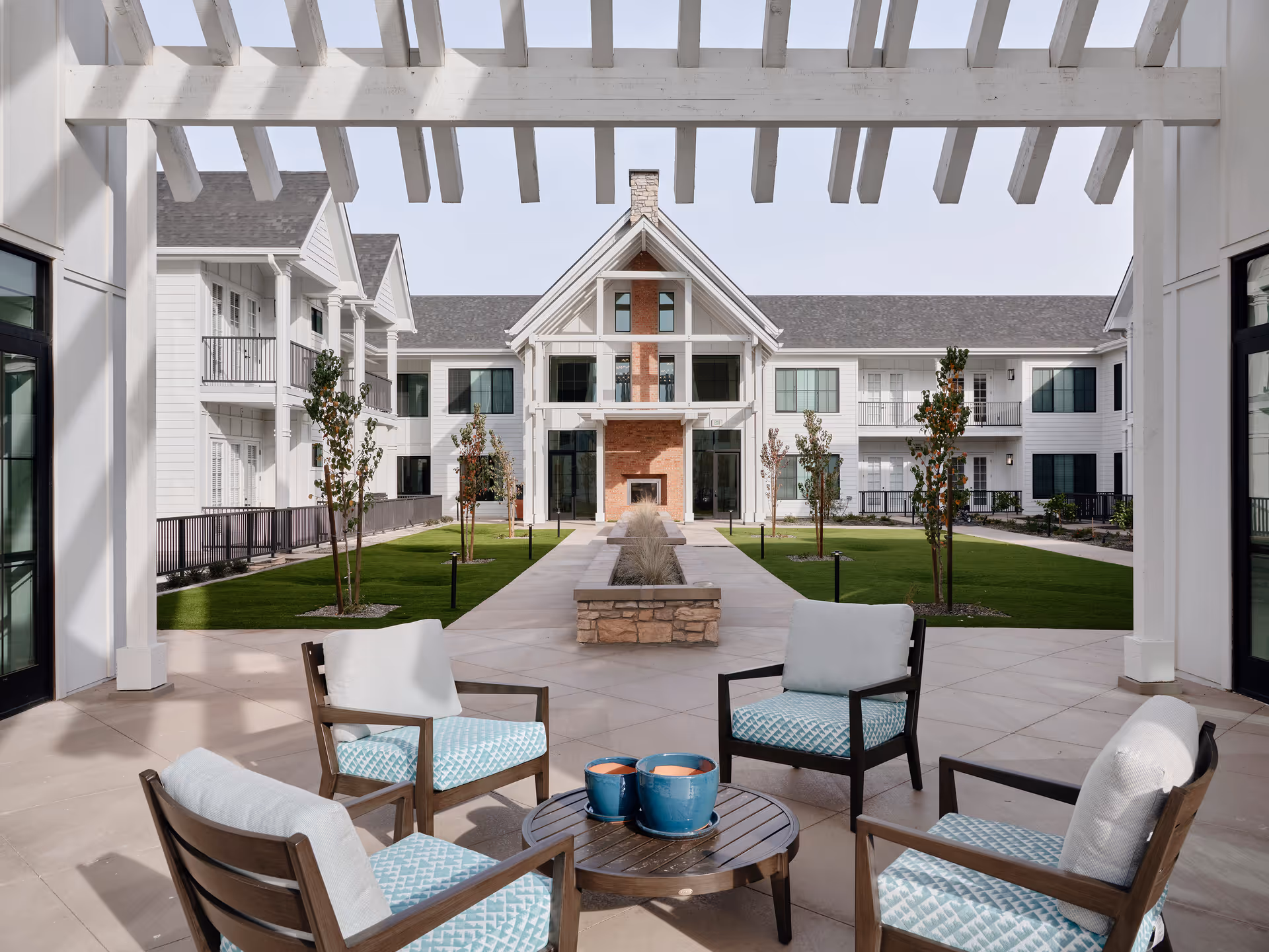 Covered courtyard with four cushioned chairs around a small table under a pergola, facing a central walkway, lawn, and a multi-story white senior living building.