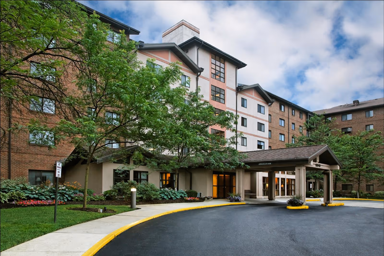 Exterior view of One Lincoln Park senior living facility showing a multi-story brick and stucco building with a covered entrance driveway, surrounded by trees and landscaped greenery under a partly cloudy sky.