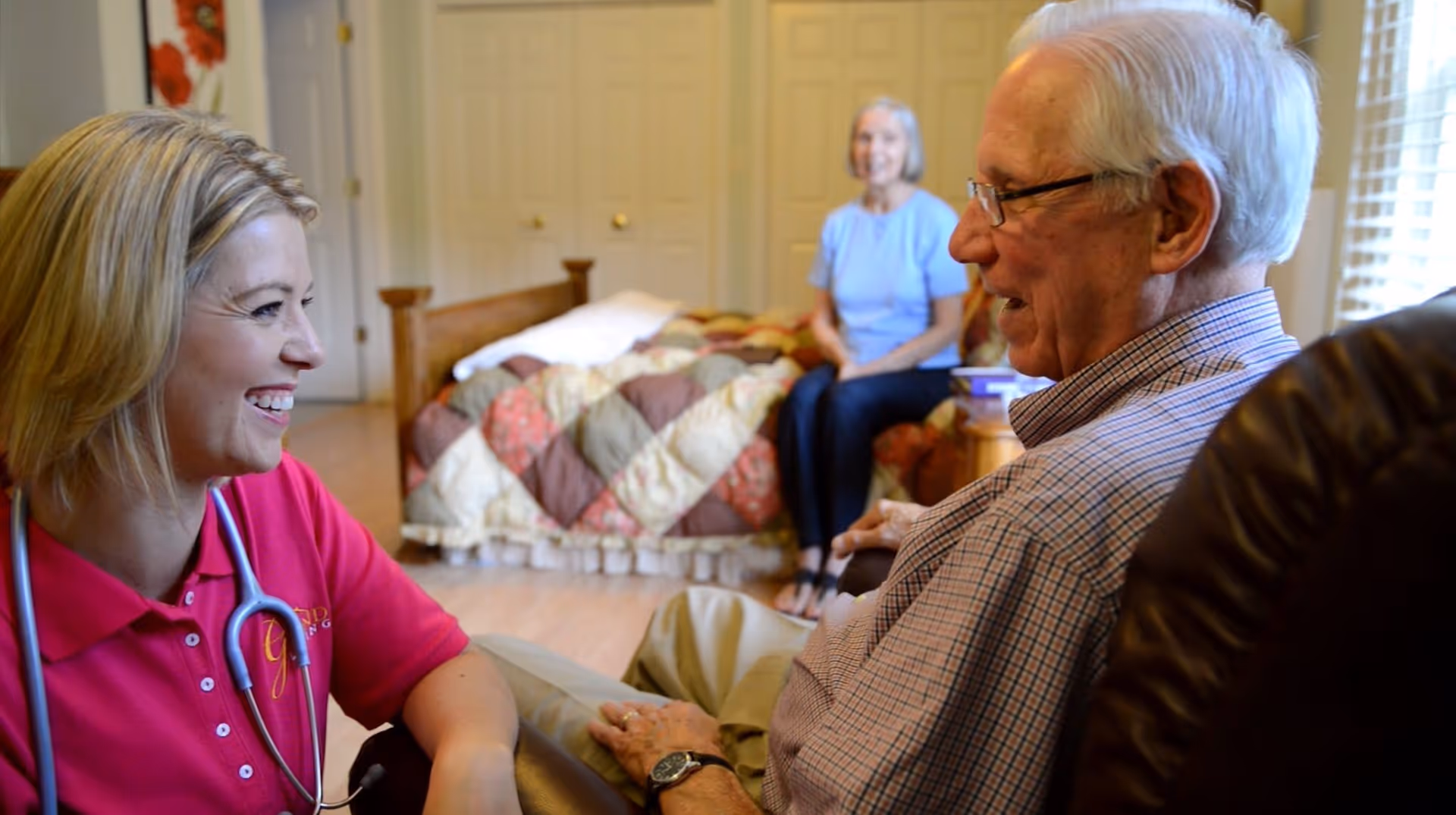 A smiling female caregiver wearing a pink shirt and stethoscope talks with an elderly man seated in a chair. In the background, an elderly woman sits on a bed with a colorful quilt in a well-lit bedroom.