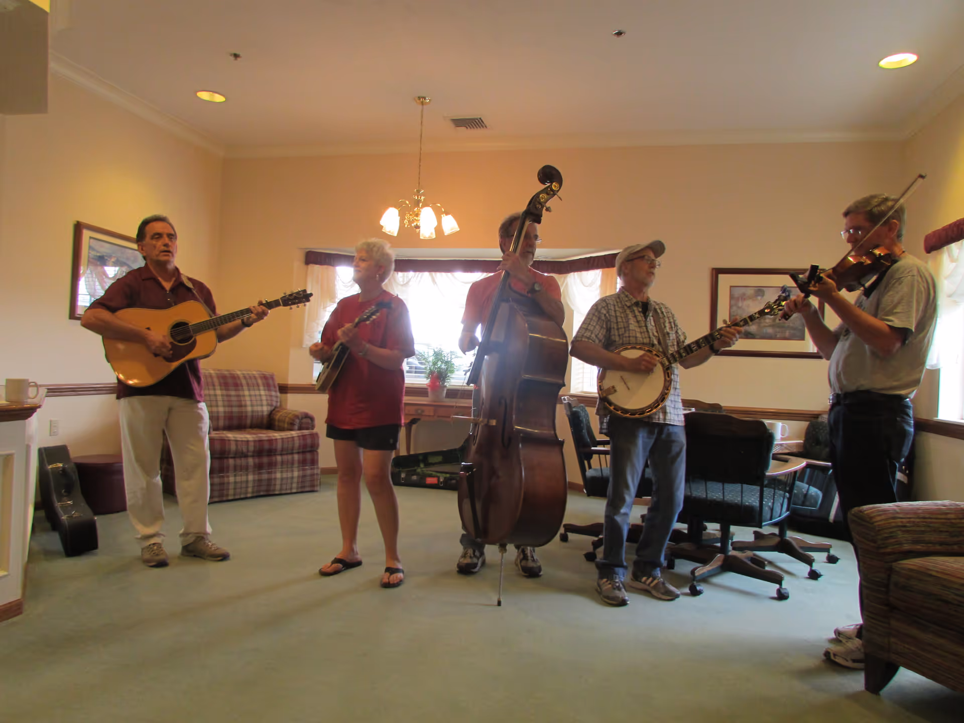 Five people playing musical instruments in a living room setting. The group includes a guitarist, a mandolin player, a double bass player, a banjo player, and a violinist. The room has beige walls, a green carpet, a plaid armchair, and a table with chairs near a window with white curtains.