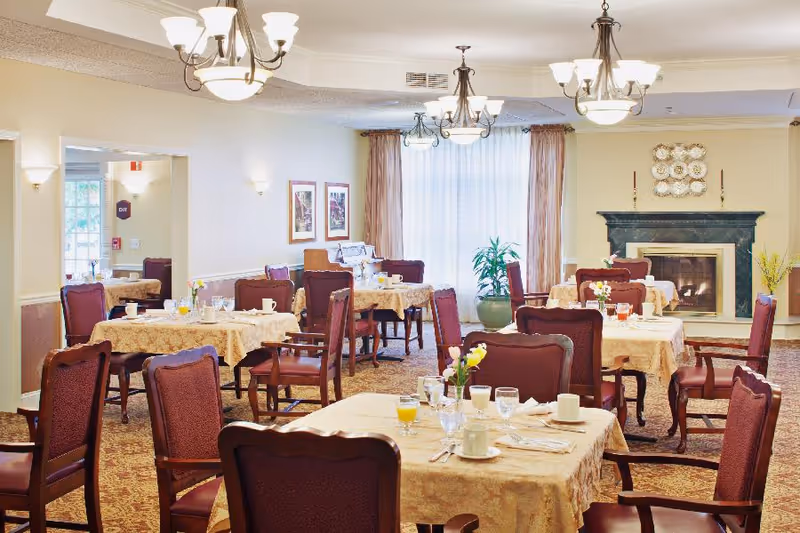 A dining room in a senior living facility with multiple tables covered in beige tablecloths, each set with cups, glasses, and silverware. The room features burgundy upholstered chairs, a fireplace with decorative plates above it, large windows with sheer curtains, and warm lighting from chandeliers and wall sconces.