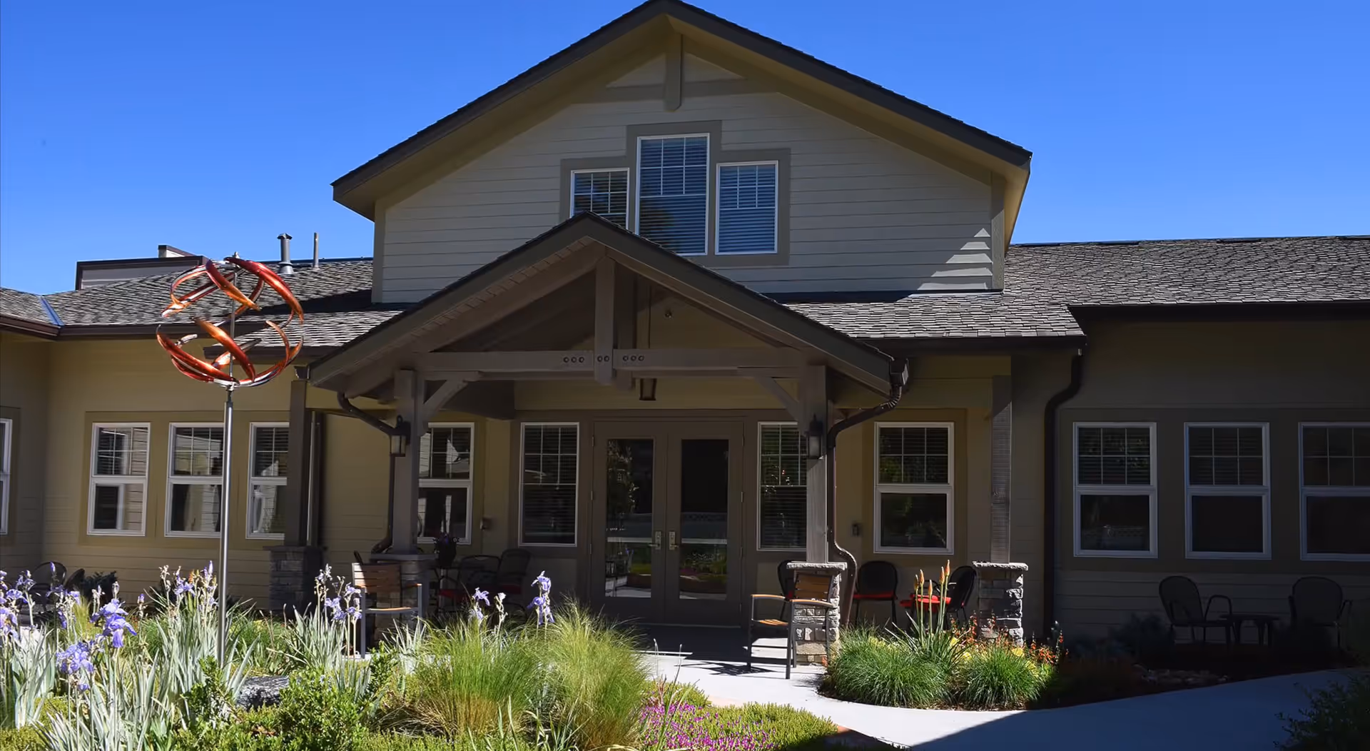 Exterior view of a senior living facility building with a covered entrance, multiple windows, outdoor chairs, and a garden with flowers and ornamental grass under a clear blue sky.