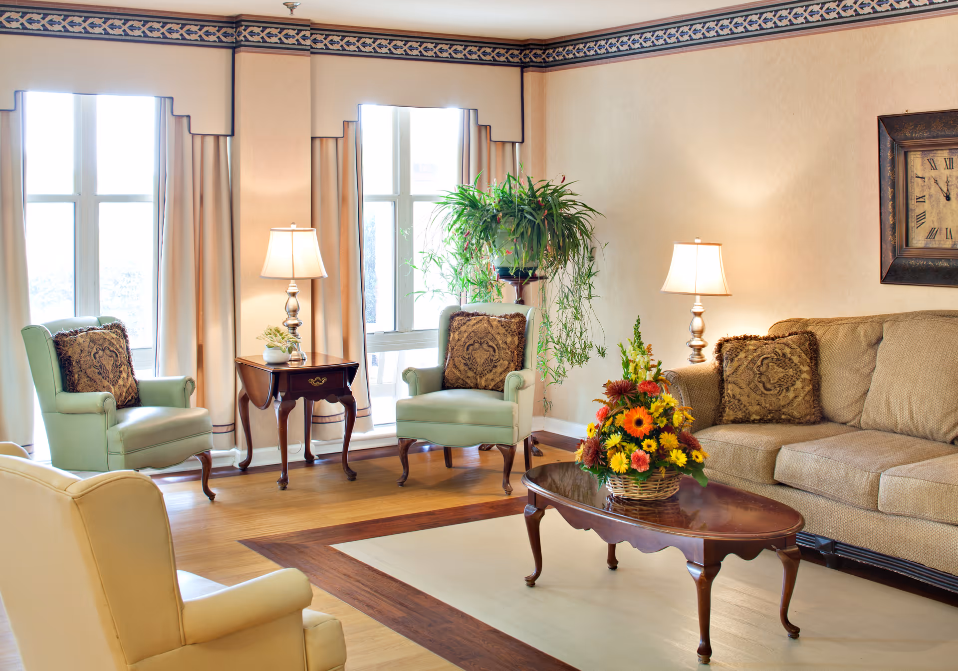 A bright, well-furnished living room with a sofa and armchairs surrounding a wooden coffee table topped with a floral arrangement, side tables with lamps, and a potted plant.