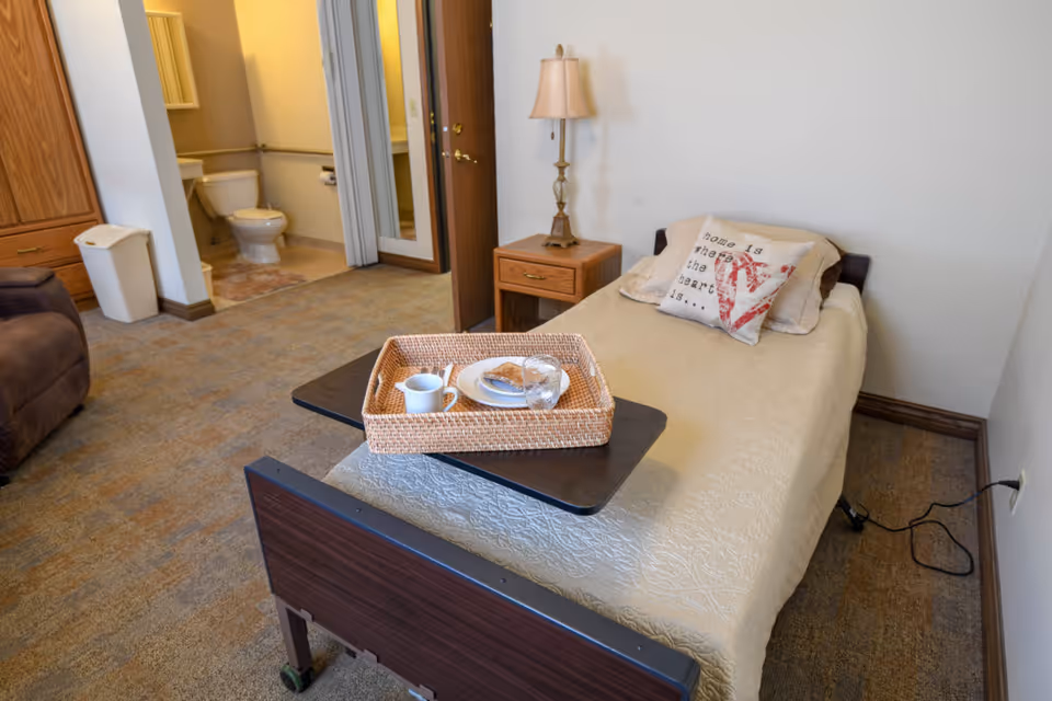 A single bed with a beige quilt and decorative pillows, including one with the text 'home is where the heart is,' is positioned in a room with carpeted flooring. A wooden nightstand with a lamp sits beside the bed. A tray with a cup, plate, and glass is placed on a movable overbed table. In the background, there is an open door leading to a bathroom with a toilet and sink visible. A brown recliner chair and a trash bin are also partially visible.