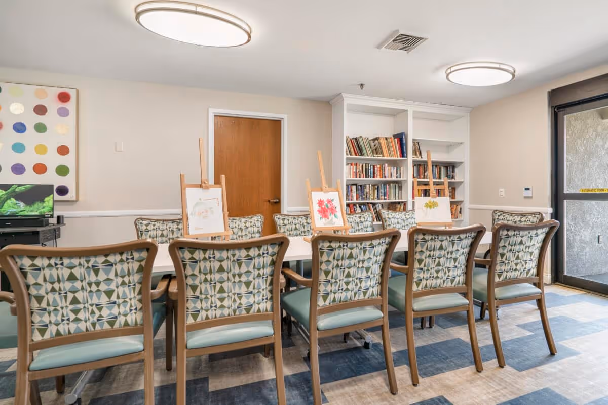 A well-lit room with a long table surrounded by patterned chairs. On the table are three easels displaying watercolor paintings of flowers. Behind the table is a white bookshelf filled with books. A wooden door and a glass door leading outside are visible in the background. The room has modern ceiling lights and a colorful abstract painting on one wall.