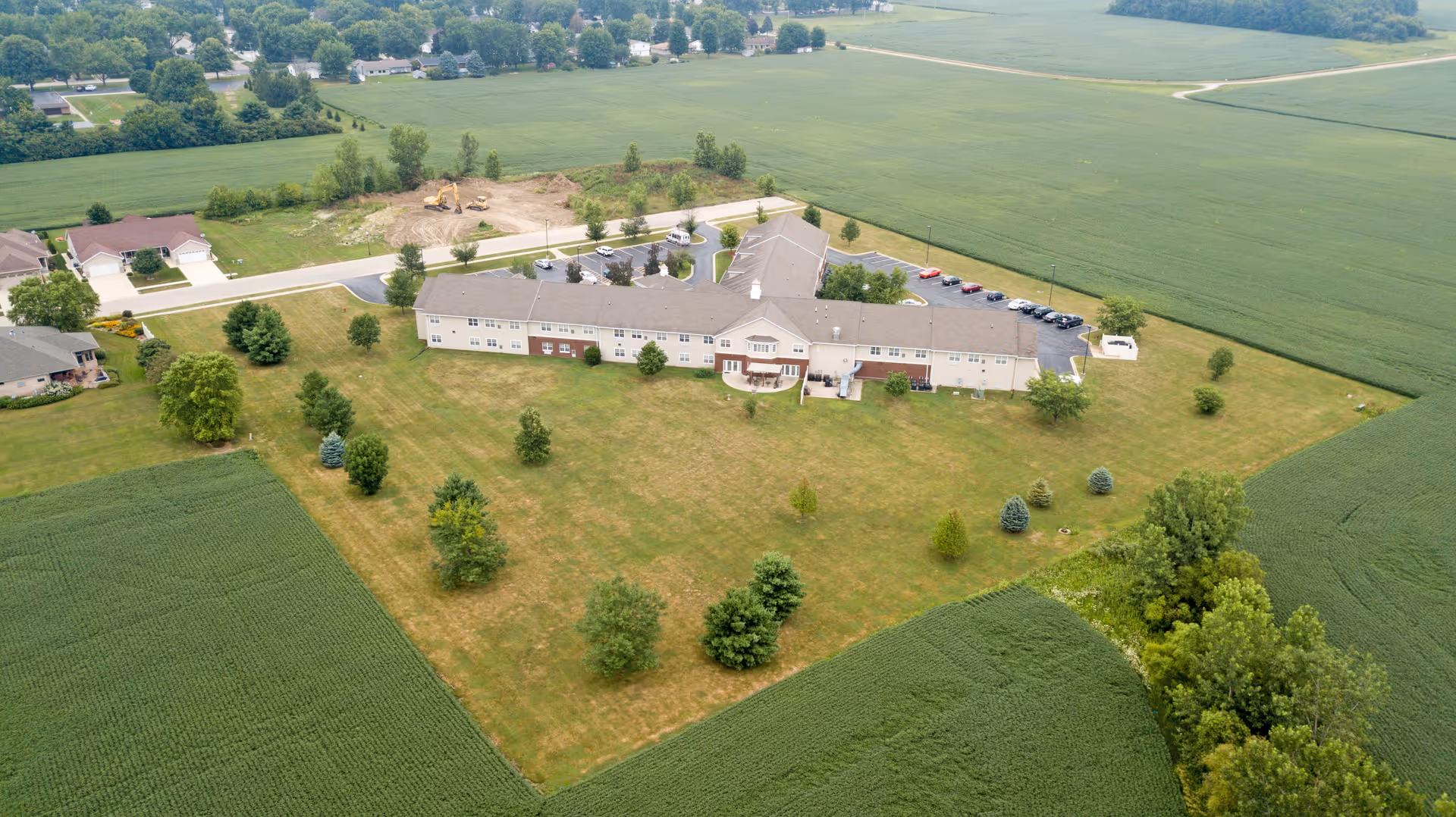 Aerial view of a long one-story senior living facility building surrounded by lawns, trees, a parking lot, and surrounding farmland.