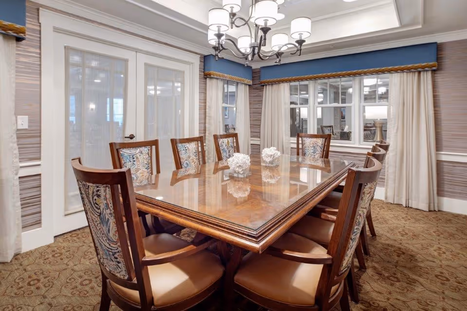 A formal dining room with a large wooden table surrounded by eight upholstered chairs. The table has a polished surface with decorative white coral-like centerpieces. The room features patterned carpet, beige curtains, blue valances, and a chandelier hanging from the ceiling. French doors and windows with white frames are visible in the background.