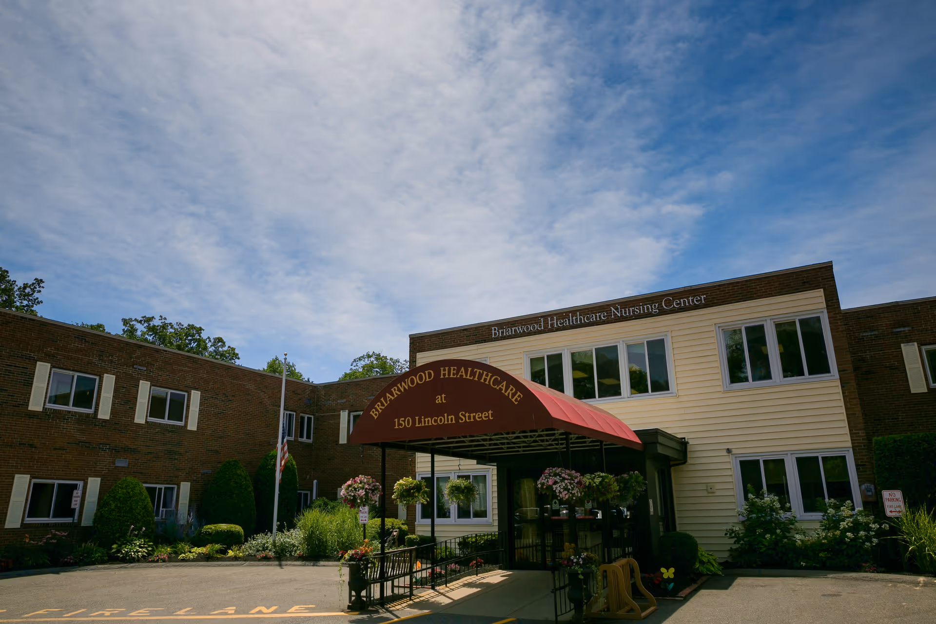 Exterior view of Briarwood Healthcare Nursing Center building with a red canopy entrance that reads 'Briarwood Healthcare at 150 Lincoln Street'. The building is two stories with brick and light-colored siding, surrounded by greenery and flowers under a partly cloudy sky.