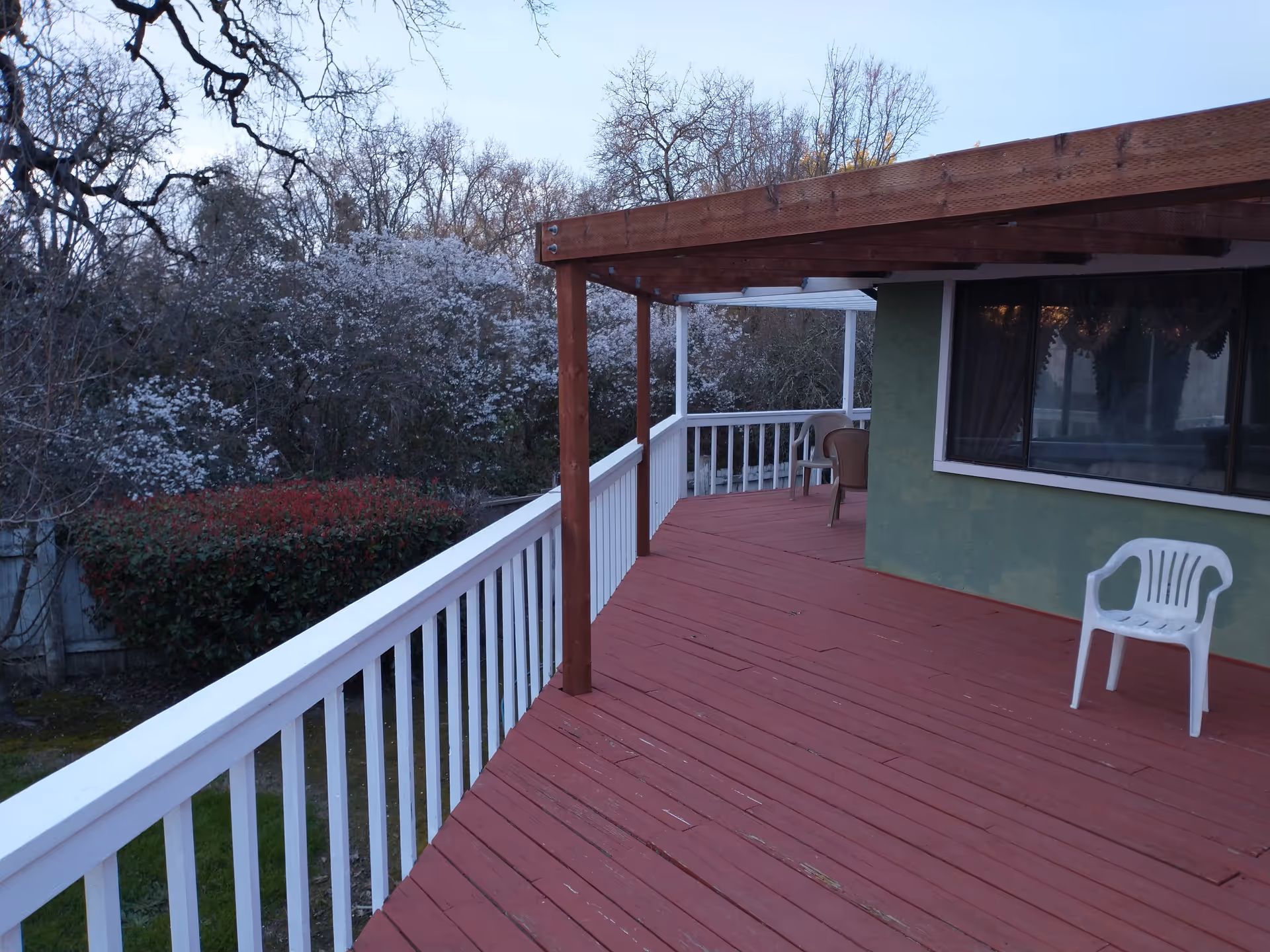 Outdoor wooden deck with red flooring and white railing attached to a green building. There is a white plastic chair on the deck and some brown chairs under a wooden pergola. Trees and bushes are visible in the background.