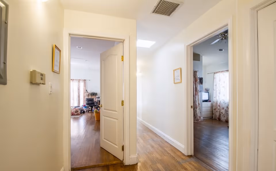A hallway with wooden flooring and cream-colored walls leading to two rooms. The room on the left has an office chair, a desk, and a window with floral curtains. The room on the right has a dresser, a television, and a window with floral curtains. There are framed pictures on the hallway walls and a ceiling vent above.