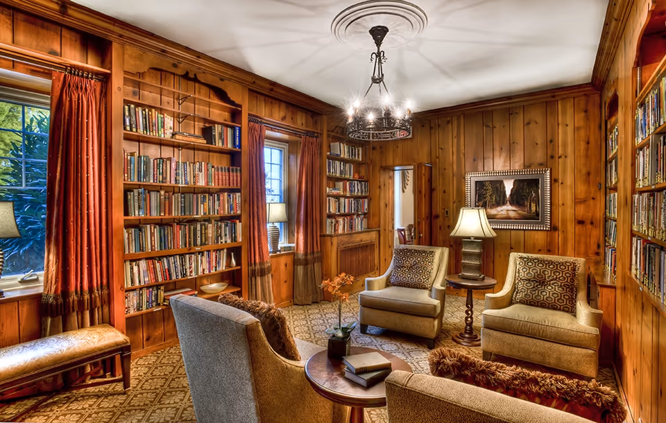 A cozy library room with wooden paneled walls and built-in bookshelves filled with books. The room features four upholstered armchairs arranged around a small round wooden table with books and a plant on it. There are two windows with red curtains, a bench under one window, a table lamp on a side table, a framed painting on the wall, and a decorative chandelier hanging from the ceiling.
