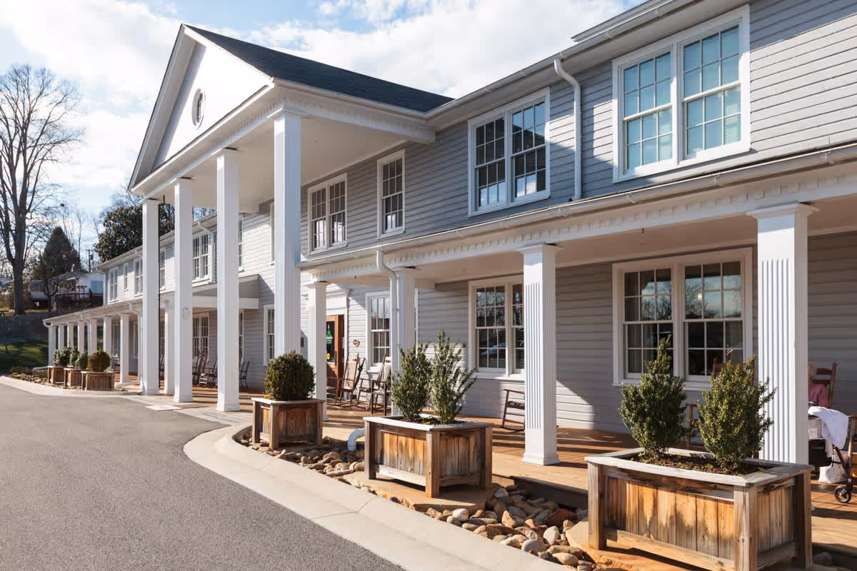 Exterior view of a two-story building with white columns and a covered porch. The building has multiple windows and wooden planters with green shrubs along the porch. There are rocking chairs on the porch and a paved driveway in front.
