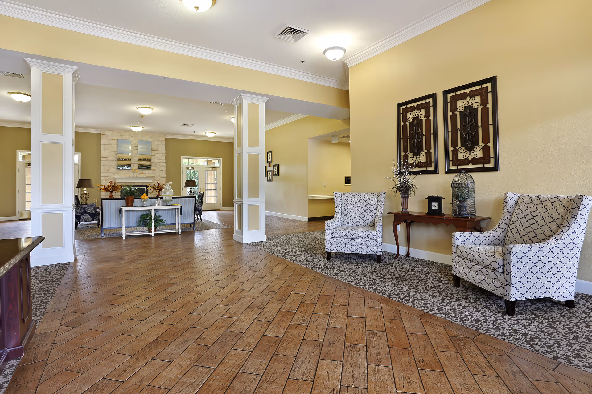 A spacious and well-lit senior living facility common area with wooden tile flooring and patterned carpet. The room features two patterned armchairs with a wooden side table between them, decorated with a vase, a clock, and a decorative birdcage. In the background, there is a seating area with a sofa, chairs, a fireplace with a stone surround, and wall art above it. The walls are painted a warm beige color, and the ceiling has recessed lighting and crown molding.