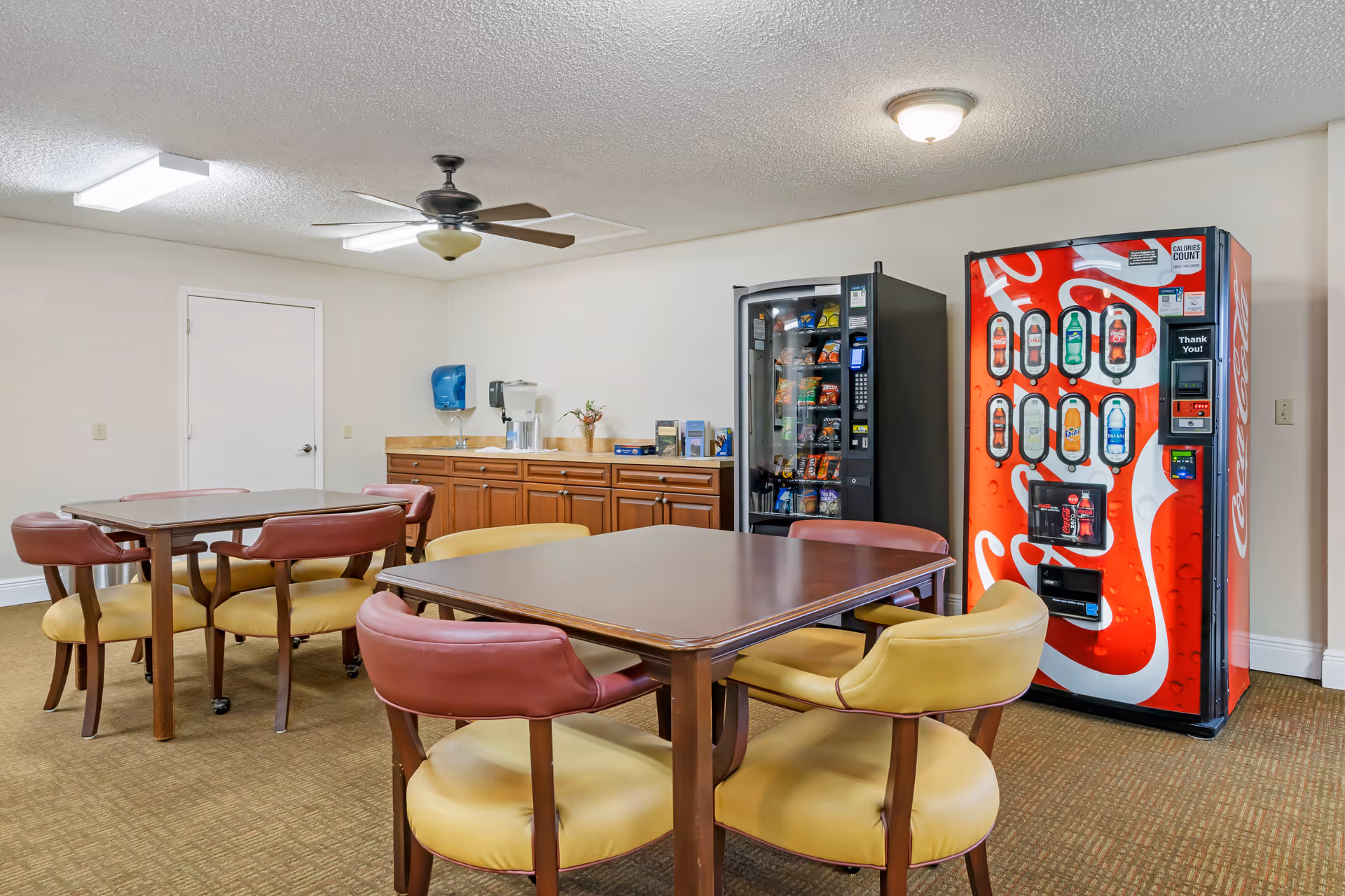 A small common area with two wooden tables surrounded by chairs with yellow seats and maroon backs. In the background, there is a countertop with a sink, a water dispenser, and some brochures. To the right, there is a vending machine stocked with snacks and a Coca-Cola beverage vending machine. The ceiling has a fan and light fixtures.