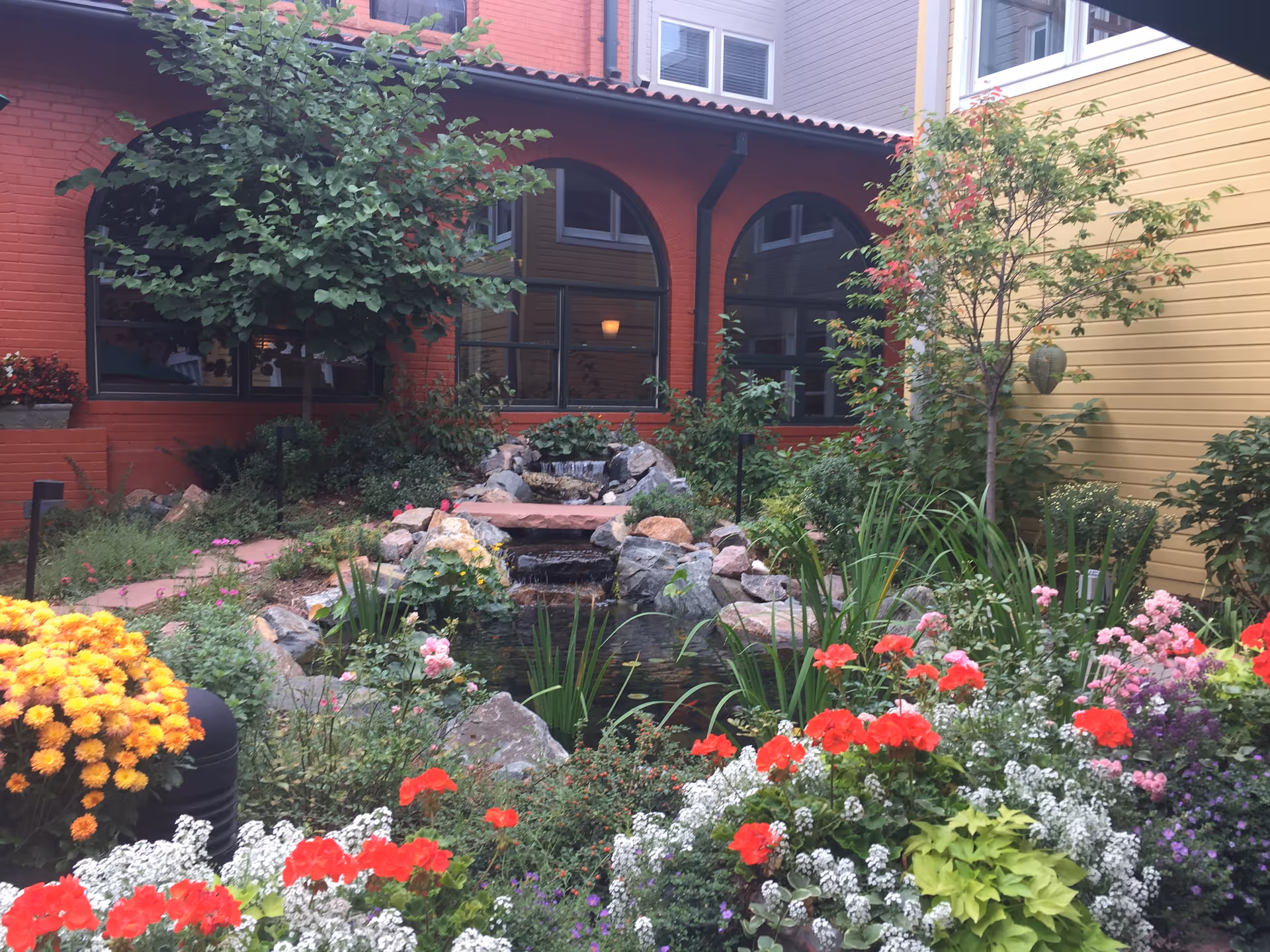 Landscaped courtyard with a small pond and waterfall surrounded by colorful flowers and arched windows of a brick building.