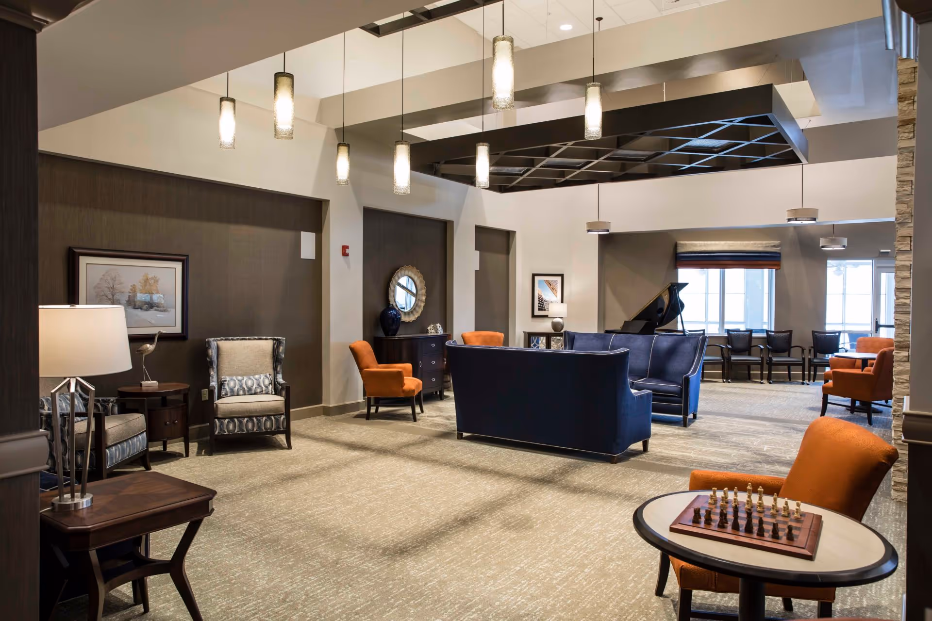 Spacious furnished communal lounge with blue sofas, orange armchairs, pendant lighting, a grand piano, and a chess set on a small table.