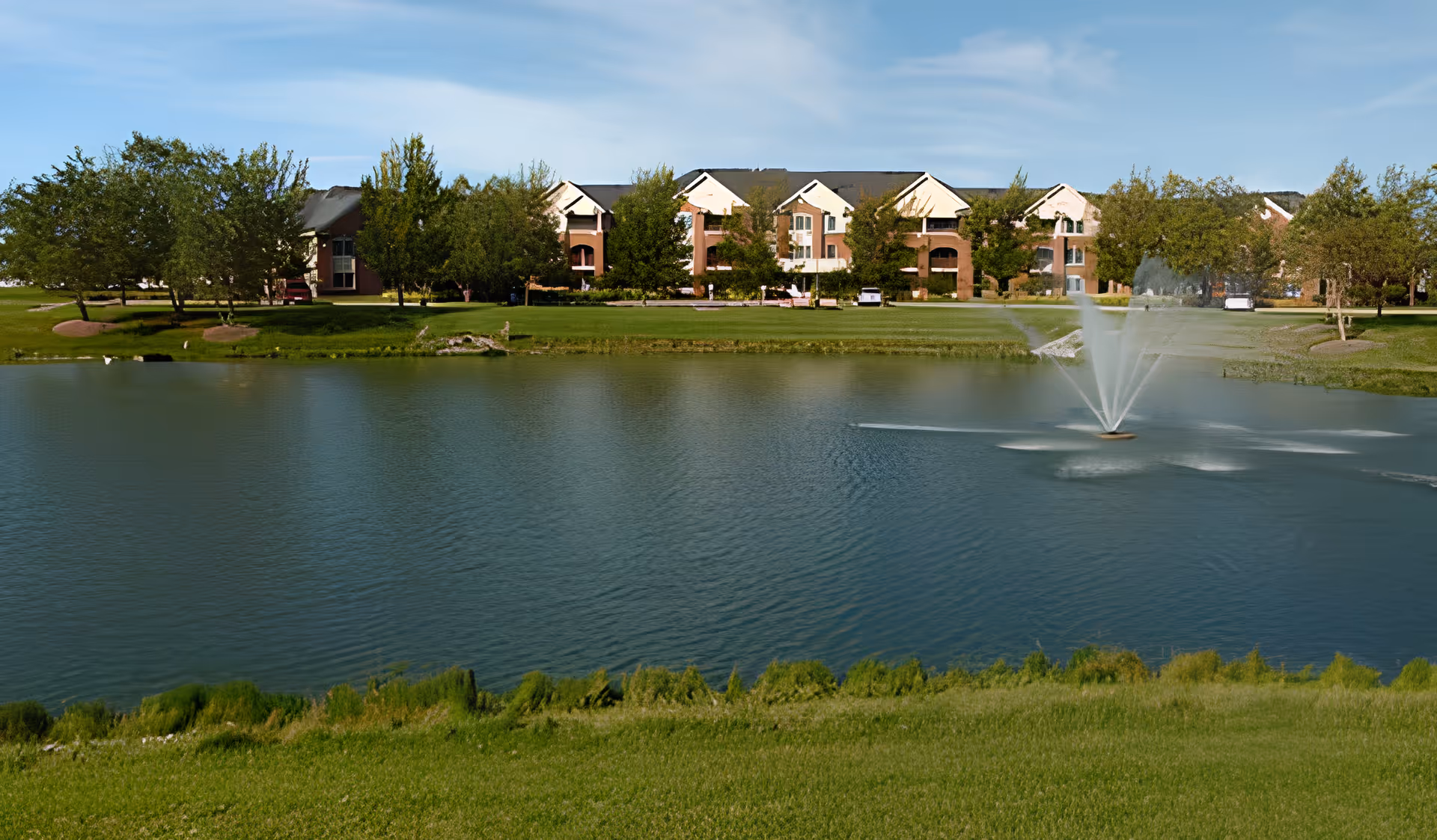 Pond with a central fountain in front of a multi-story assisted living building surrounded by trees and lawn.